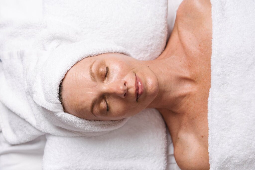 A top-down view of a woman lying down with her eyes closed, relaxing during a spa treatment. Her head rests on a pillow with a white towel wrapped around her hair, and her chest is covered by a white towel.