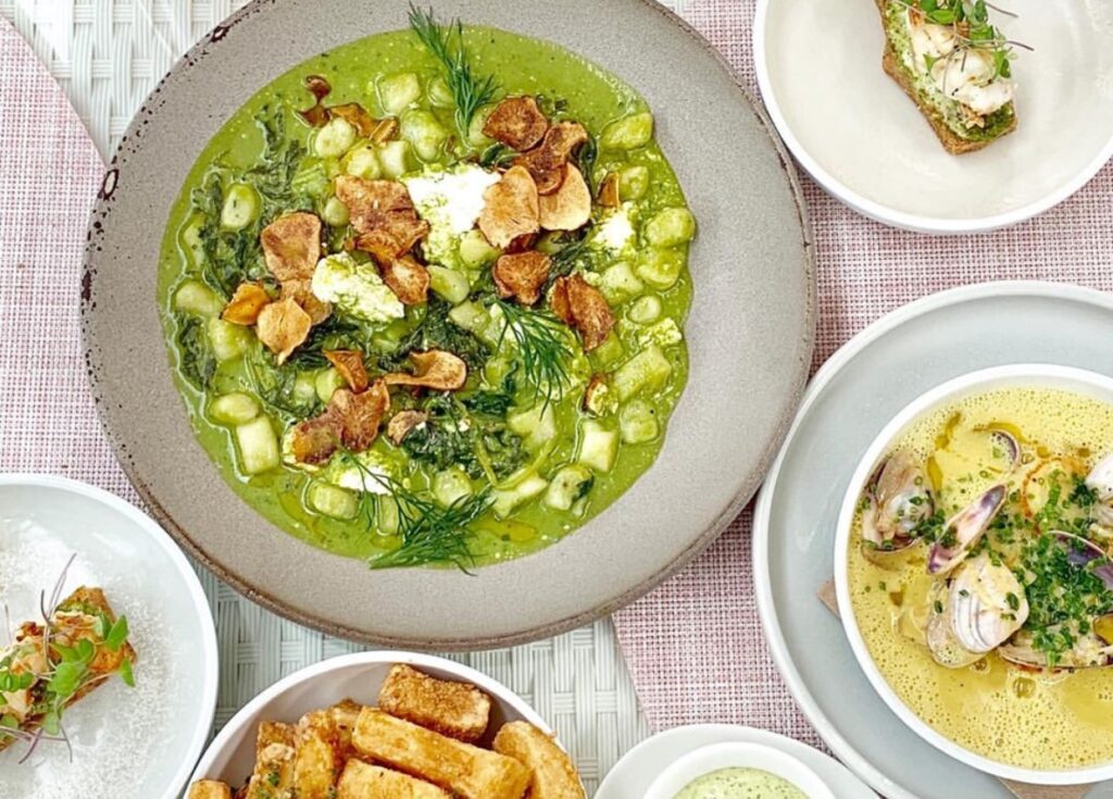 A top-down view of a dining table featuring a variety of plated dishes. The main dish on the left is a gnocchi plate served in a vibrant green sauce.