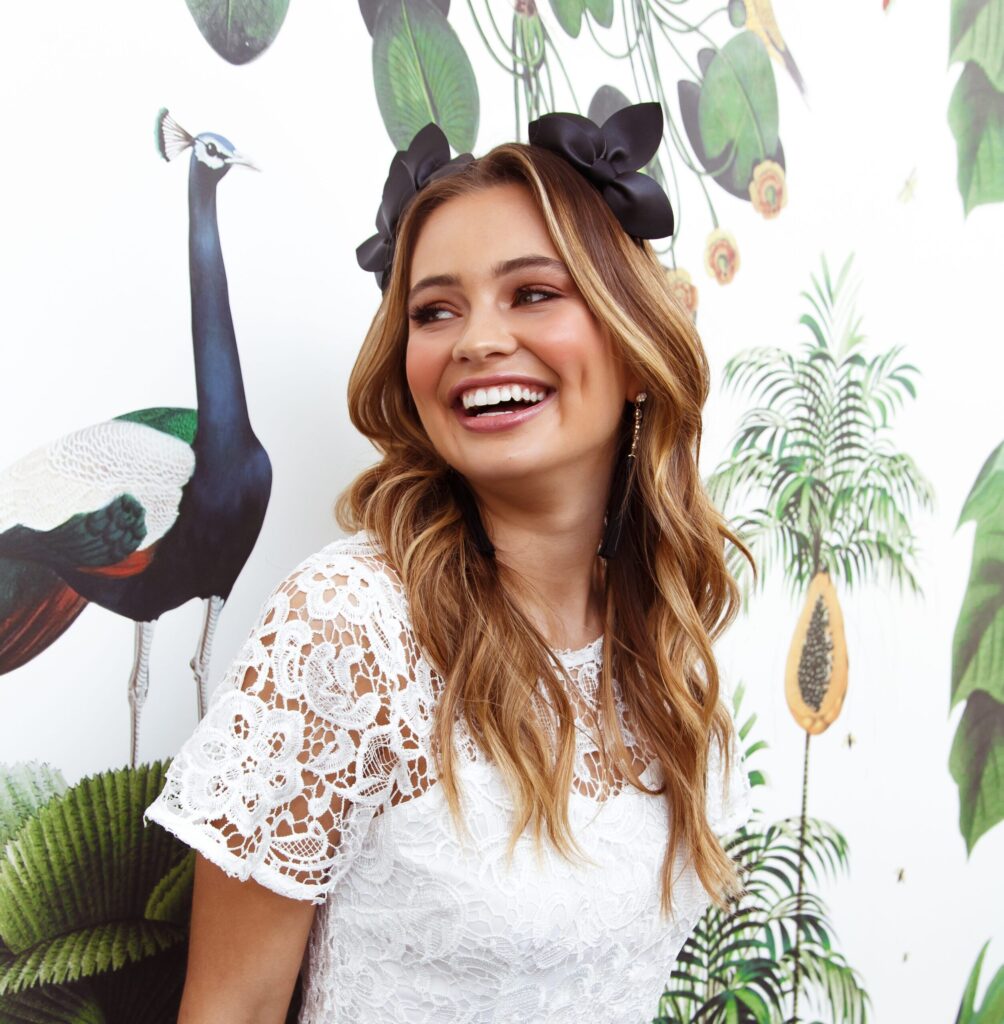 A smiling woman with wavy blonde hair and a black bow headband poses in a white lace dress against a wallpaper background featuring a peacock and tropical plants.