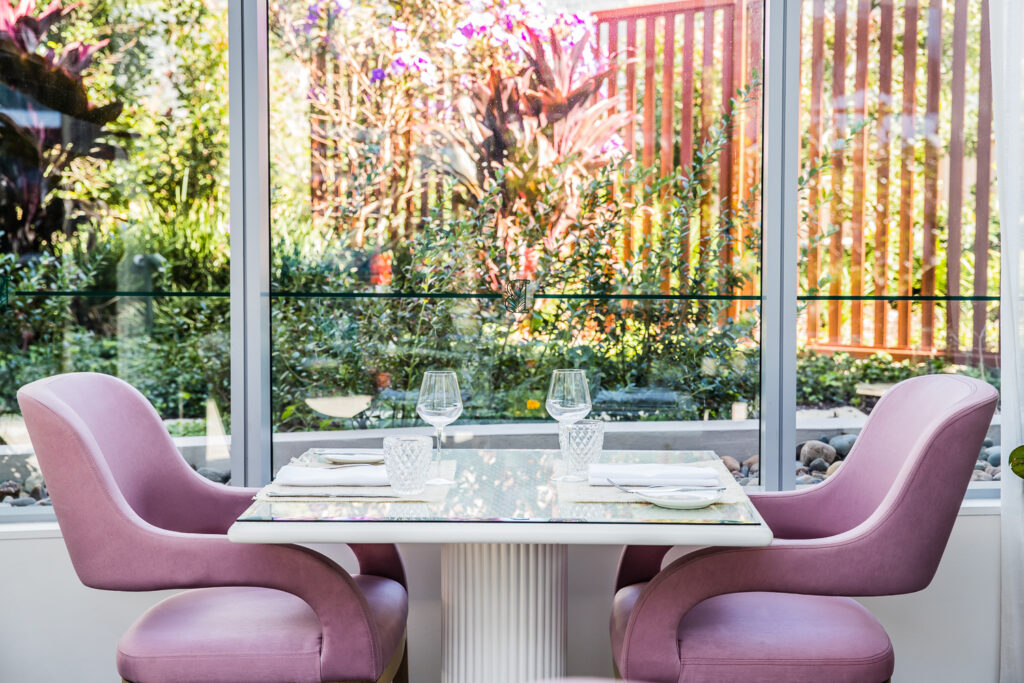 An elegant table set for two with wine glasses and white napkins, flanked by two plush pink armchairs. The table sits in front of a large glass window overlooking a sunlit garden with green foliage and a wooden fence.