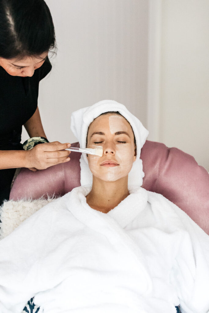 A Sol Spa professional applies a white facial mask with a brush to a client who is relaxing in a plush pink chair while wearing a white bathrobe and head towel.