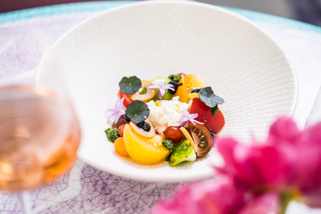A close-up shot of a colorful salad served in a large white bowl, featuring sliced red and yellow tomatoes, green herbs, a central scoop of soft white cheese, and tiny purple flowers.