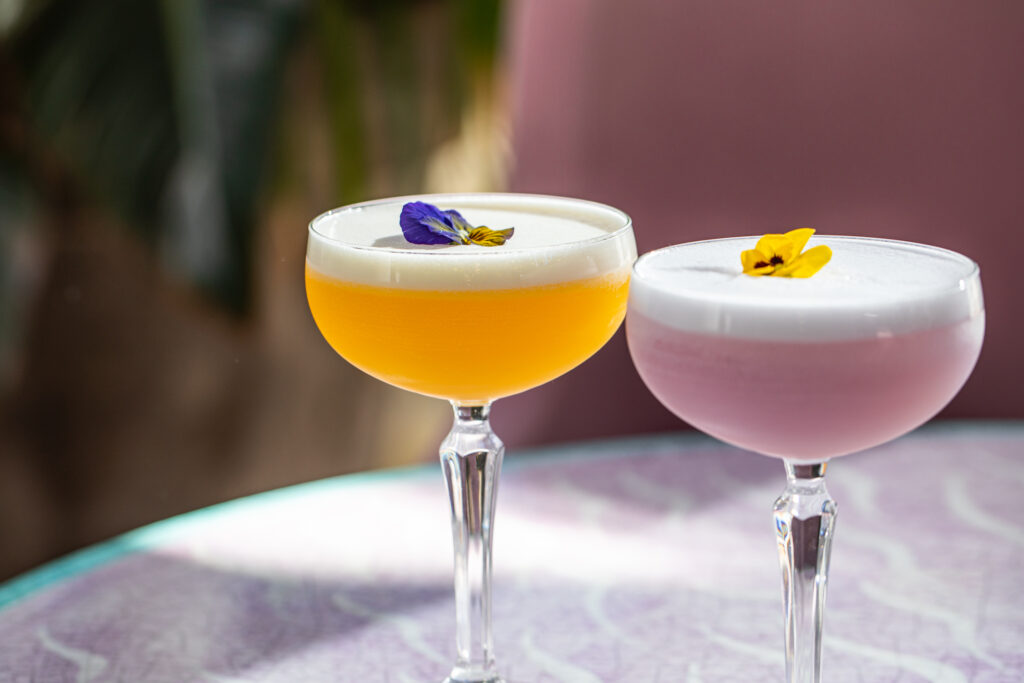 Two vibrant cocktails served in coupe glasses sit on a patterned table. The left drink is a bright yellow-orange color topped with white foam and a purple pansy, while the right drink is a pastel pink color topped with white foam and a yellow pansy.