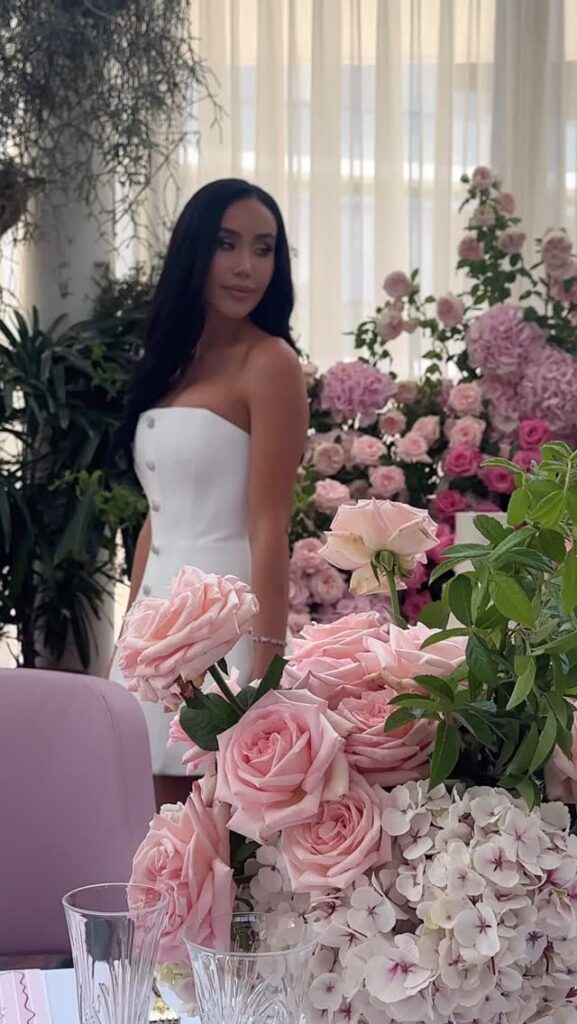 A woman in a strapless white dress stands amidst a lavish arrangement of pink roses and hydrangeas in an elegant botanica.