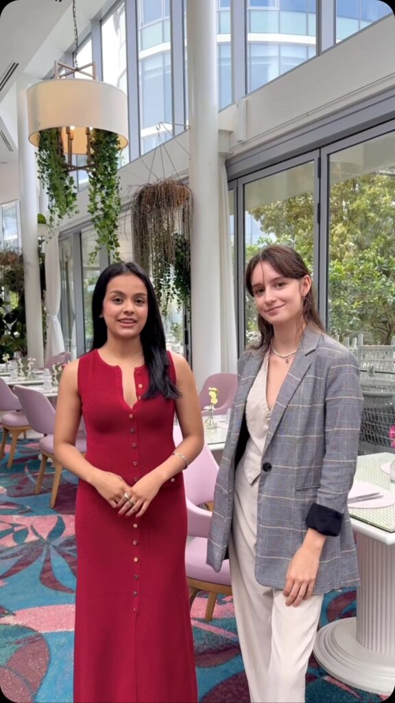 Two women stand side-by-side inside a bright, modern restaurant with high ceilings and large windows. The woman on the left wears a sleeveless, button-down red dress, while the woman on the right wears a grey plaid blazer over a light-colored vest and pants.